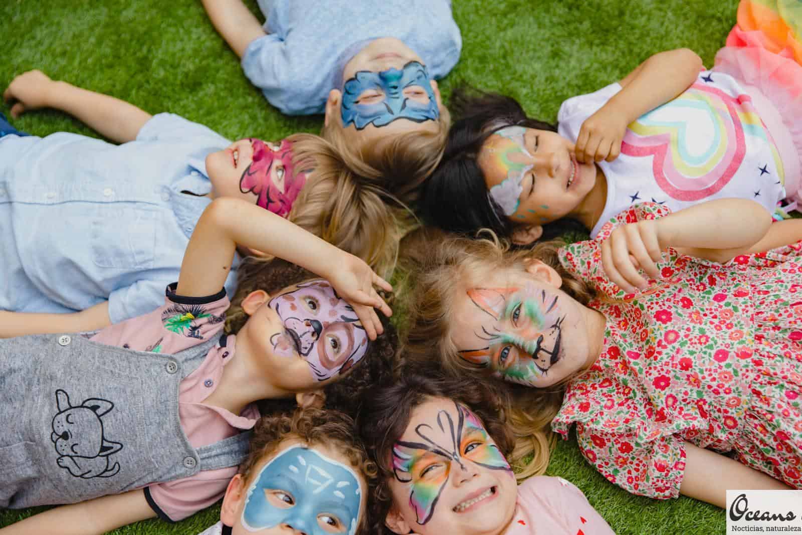 Group of children enjoying an outdoor party with colorful face paint, lying on grass.